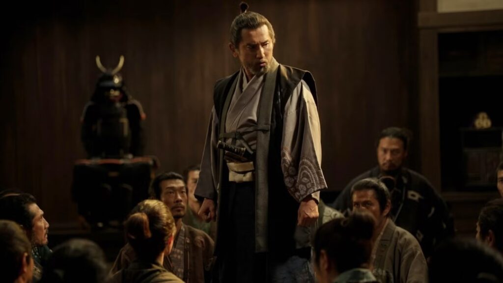 a Bearded Man in Traditional Samurai Robes Stands Above a Seated Crowd in a Dim Wooden Room with a Helmeted Armor Display to His Left