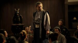 a Bearded Man in Traditional Samurai Robes Stands Above a Seated Crowd in a Dim Wooden Room with a Helmeted Armor Display to His Left