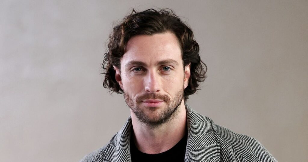 Portrait of a Man with Wavy Brown Hair and Light Beard Wearing a Gray Herringbone Coat Against a Neutral Background