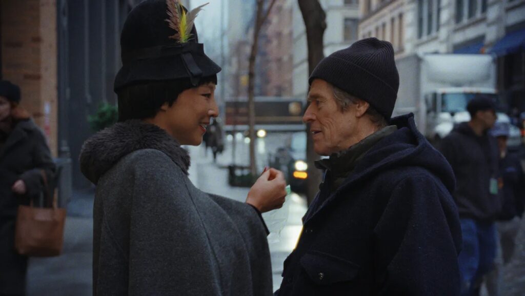Two People Converse on a City Sidewalk a Smiling Woman in a Dark Coat with a Feathered Hat Talks to a Man in a Beanie and Dark Jacket