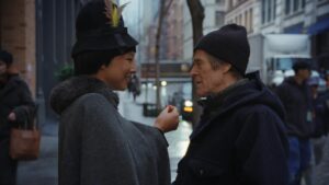 Two People Converse on a City Sidewalk a Smiling Woman in a Dark Coat with a Feathered Hat Talks to a Man in a Beanie and Dark Jacket