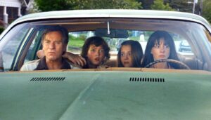 a Family of Four Sits in a Vintage Green Car the Parents in the Front Seats and Two Kids in the Back All Looking Tense As They Drive Down the Street Front View