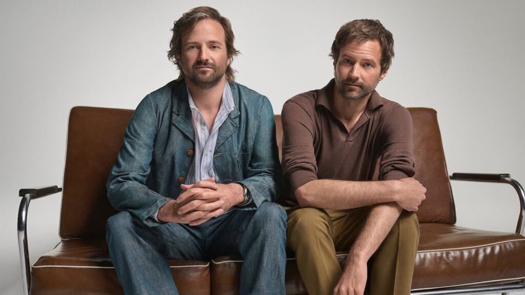 Two Men Sit Side by Side on a Brown Leather Sofa in a Studio Setting Looking at the Camera with Relaxed Expressions