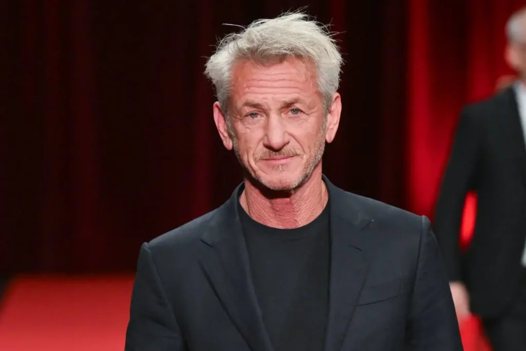 Middle aged Man with Silver Hair and Light Stubble Wearing a Black Blazer on a Red Carpet Backdrop
