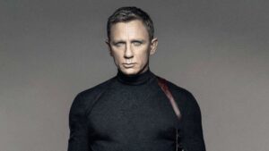 Man with a Stern Expression Wearing a Black Turtleneck and a Diagonal Leather Strap Across His Chest Against a Gray Gradient Background