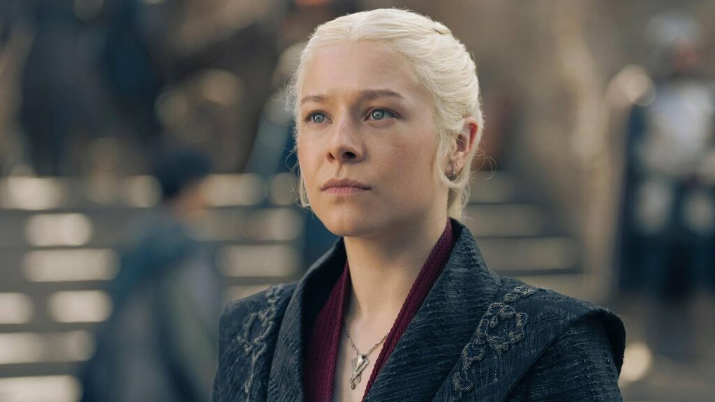 Portrait of a Thoughtful Woman with Platinum Blonde Hair Tied Back Wearing a Dark Embroidered Coat and Maroon Scarf Looking Upward in a Blurred Urban Background