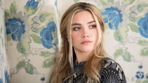 Portrait of a Woman with Long Blonde Hair Looking to the Right Against a Blue green Floral Wallpaper Backdrop and Wearing a Dark Patterned Top