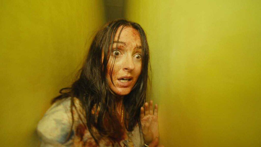 Frantic Woman with Long Dark Hair and Blood on Her Face and Clothes Pressed Against a Yellow Wall Eyes Wide in Fear