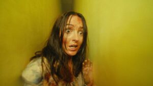 Frantic Woman with Long Dark Hair and Blood on Her Face and Clothes Pressed Against a Yellow Wall Eyes Wide in Fear