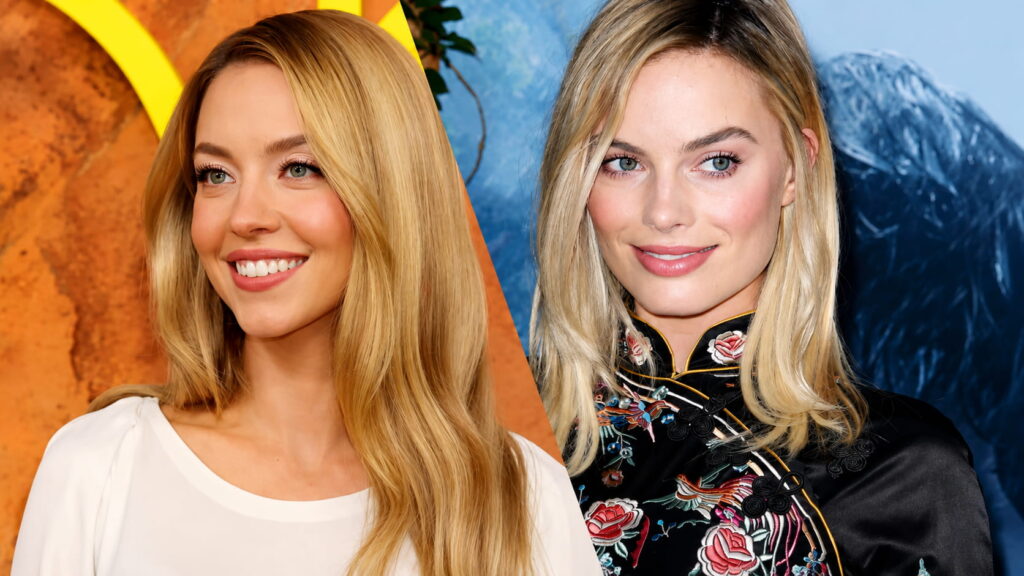 Two Blonde Women Pose Side by Side at an Event Smiling Left Wears a White Blouse Right Wears a Black Embroidered Dress Against a Colorful Backdrop