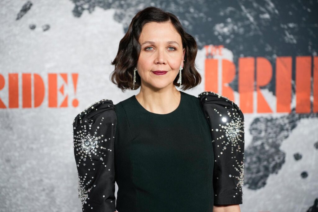 Portrait of a Woman with Shoulder length Dark Hair in a Black Dress with Jeweled Sleeves Standing Against a Backdrop with Orange Letters