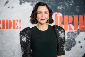 Portrait of a Woman with Shoulder length Dark Hair in a Black Dress with Jeweled Sleeves Standing Against a Backdrop with Orange Letters