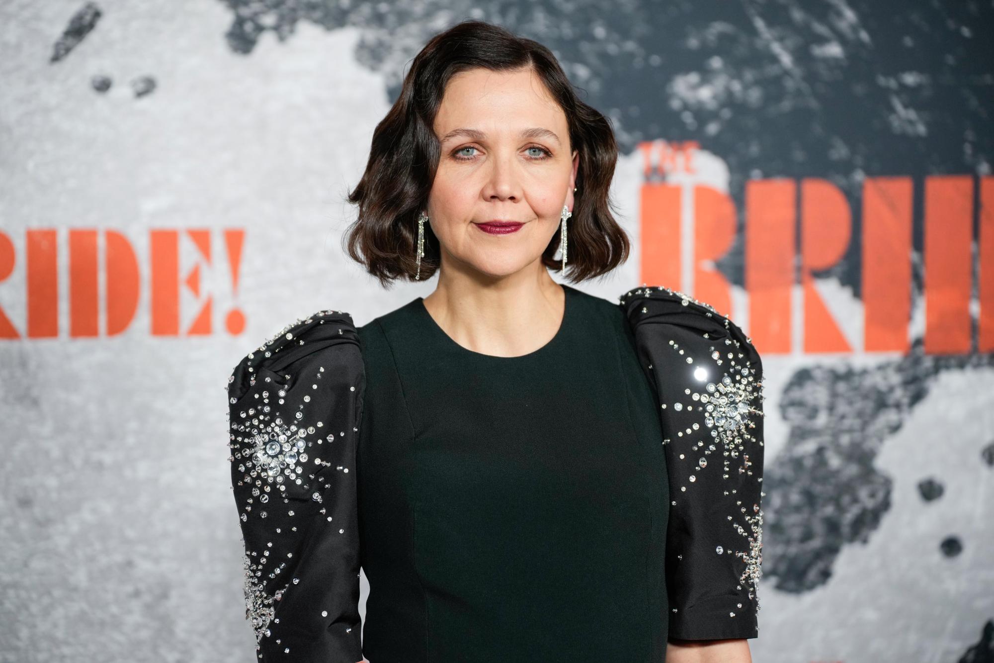 Portrait of a Woman with Shoulder length Dark Hair in a Black Dress with Jeweled Sleeves Standing Against a Backdrop with Orange Letters