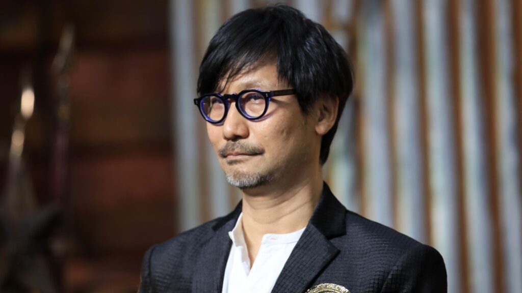 Man with Black Hair and Blue rimmed Round Glasses Wearing a White Shirt and Dark Blazer Looking to the Side