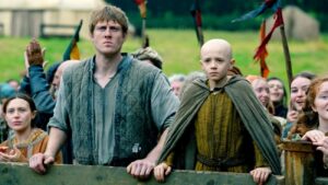 Adult Man and a Bald Boy in Medieval Costumes Grip a Wooden Railing at a Reenactment Crowd Both Looking Ahead with Serious Expressions