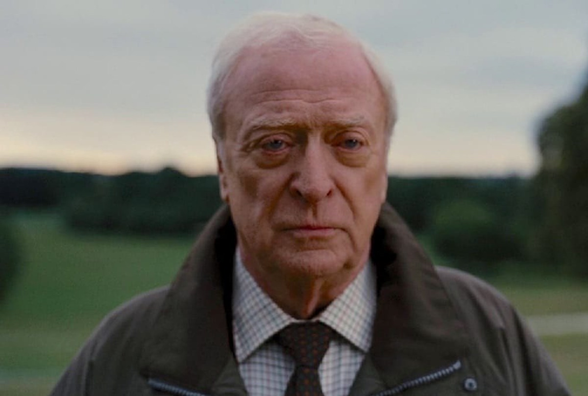 Portrait of an Elderly Man with White Hair Outdoors in a Green Field Wearing a Brown Coat and Checkered Shirt with a Brown Tie Looking Solemn