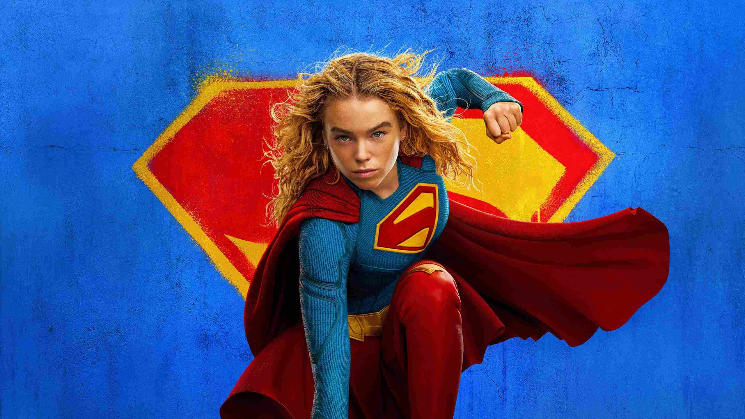 Supergirl in a Blue Suit and Red Cape Flies Forward Set Against a Blue Textured Wall with a Yellow red Shield Emblem Behind Her