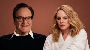 Two Adults Sit Side by Side in a Studio Setting a Man with Glasses in a Dark Sweater and White Collar and a Blond Woman in a White Blouse Against a Warm Brown Backdrop