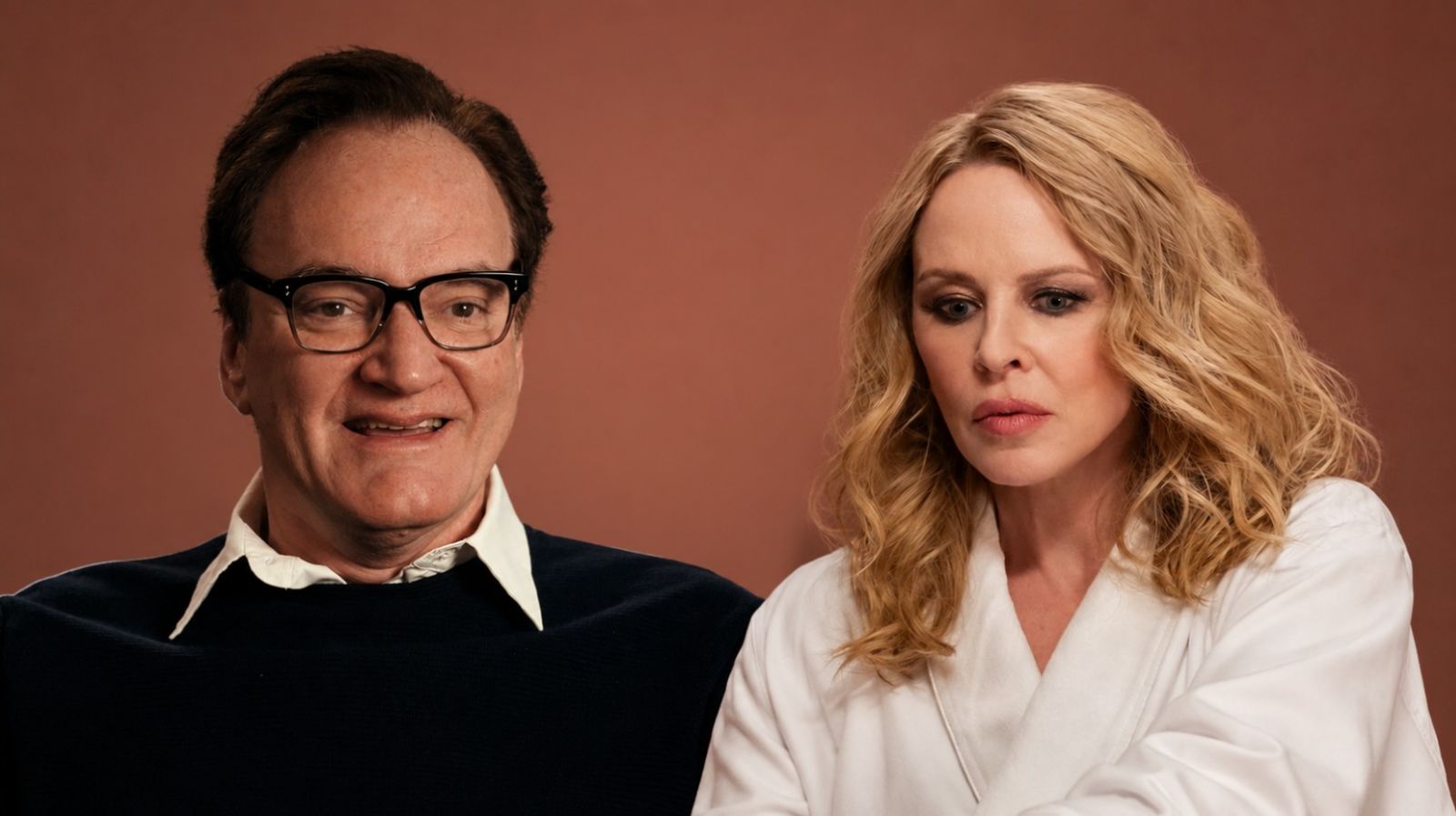 Two Adults Sit Side by Side in a Studio Setting a Man with Glasses in a Dark Sweater and White Collar and a Blond Woman in a White Blouse Against a Warm Brown Backdrop