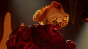 Scary Clown with Cracked Pale Makeup Red Blood Around the Mouth and Bright Orange Hair in a Red Ruffled Costume Screaming Menacingly