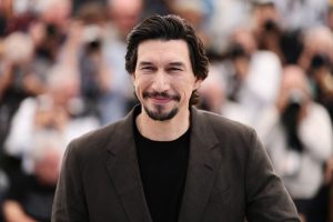 Man in a Brown Blazer and Black Shirt Smiles at the Camera with a Blurred Crowd of Photographers Behind Him