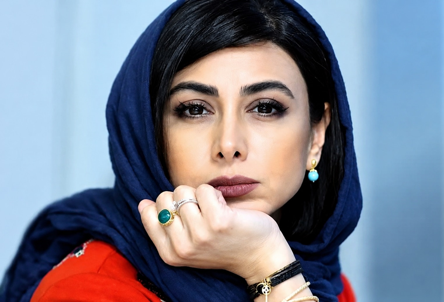 Close up of a Woman Wearing a Navy Hijab Resting Her Chin on Her Hand with Bold Makeup and Turquoise Jewelry Red Top Visible