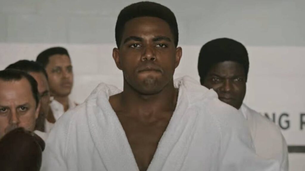 Young Black Man in a White Martial Arts Gi Walks Forward with a Serious Expression Surrounded by Onlookers in a Gym or Dojo Backdrop