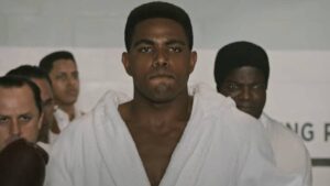 Young Black Man in a White Martial Arts Gi Walks Forward with a Serious Expression Surrounded by Onlookers in a Gym or Dojo Backdrop