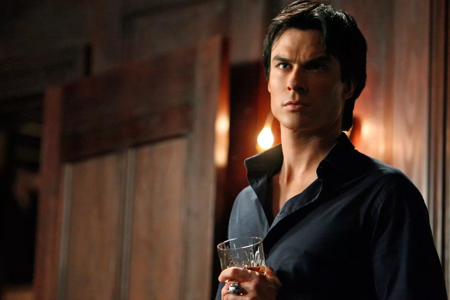 Man in a Black Shirt Stands in a Warmly Lit Wooden Bar Holding a Whiskey Glass