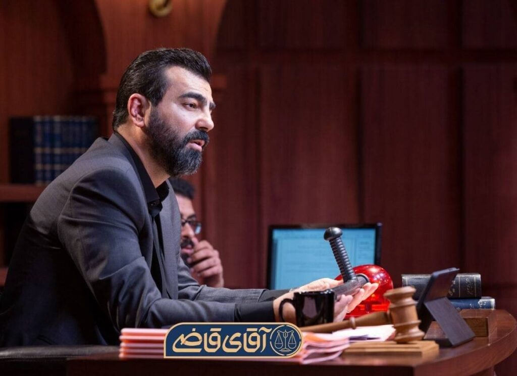 a Bearded Man in a Dark Suit Sits at a Wooden Desk with a Gavel Red Ornament and Stacks of Papers in a Courtroom or Office Setting Focusing Ahead