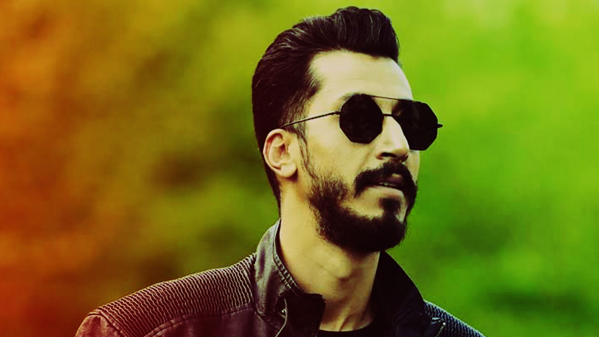 Man with a Beard and Mustache Wearing Dark Sunglasses Looking off to the Side Against a Blurred Greenorange Background