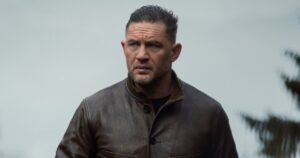 Man with Short Slicked back Hair and Stubble Wearing a Brown Leather Jacket Outdoors Against a Cloudy Sky Looking off to the Side