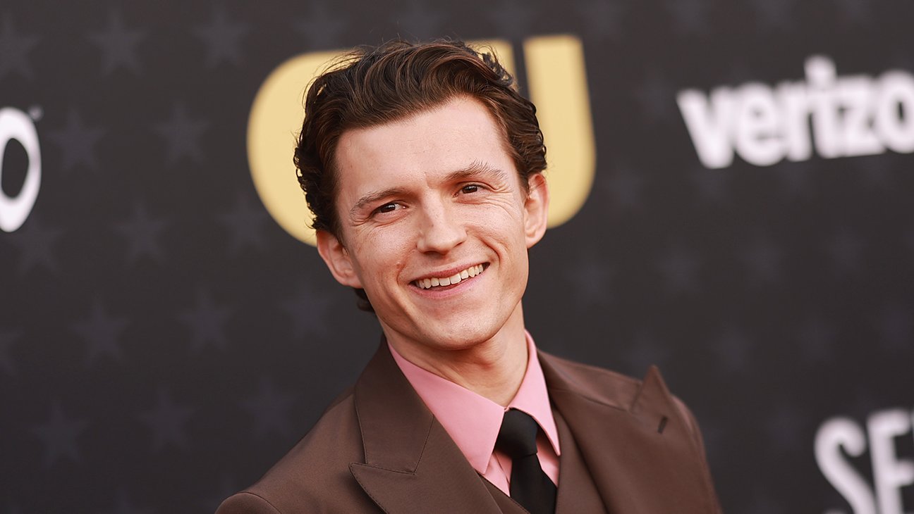 Smiling Man in a Brown Suit with a Pink Shirt and Black Tie Posing on a Red Carpet Backdrop with Logos Behind Him