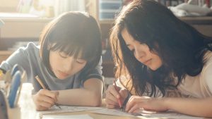 Two People Sit at a Table Drawing on Papers with Pencils in a Sunlit Room Concentrating on Their Artwork and Supplies Nearby