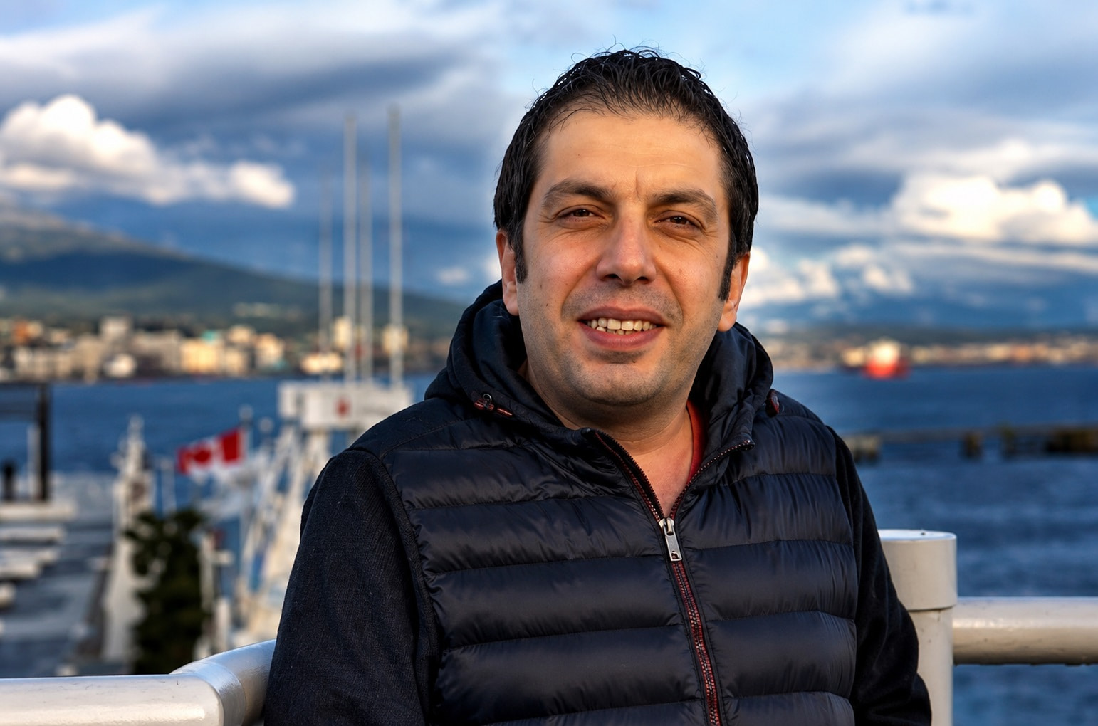 Smiling Man in a Black Quilted Vest Stands at a Harbor Railing with Water and Boats in the Background