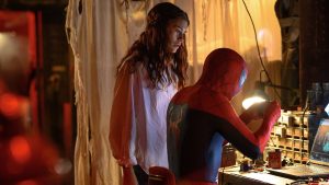 Spider man in a Red and blue Suit Leans over a Cluttered Workbench Talking to a Woman with Long Dark Hair in a Dim Warm lit Workshop