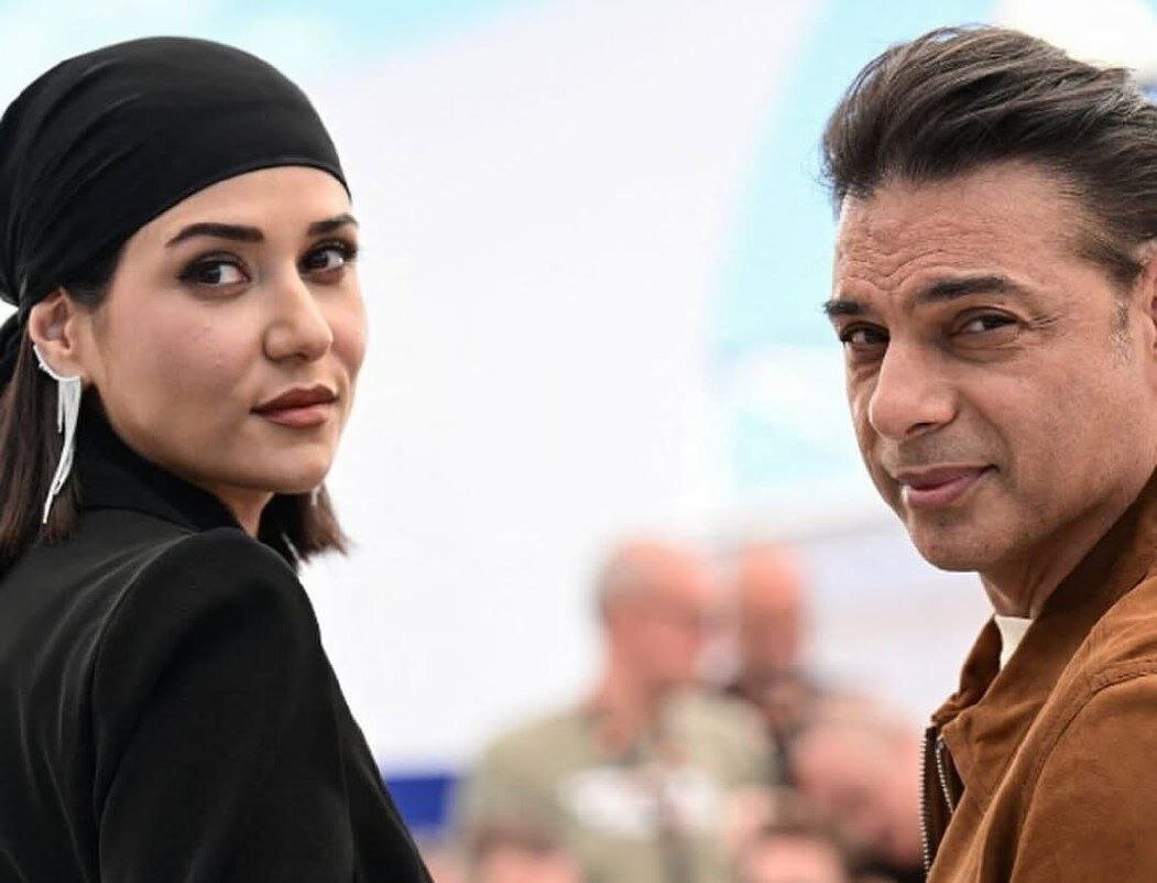 Woman in a Black Headscarf and a Man in a Brown Jacket Turn Toward the Camera in a Bright Outdoor Setting Both Looking Back over Their Shoulders