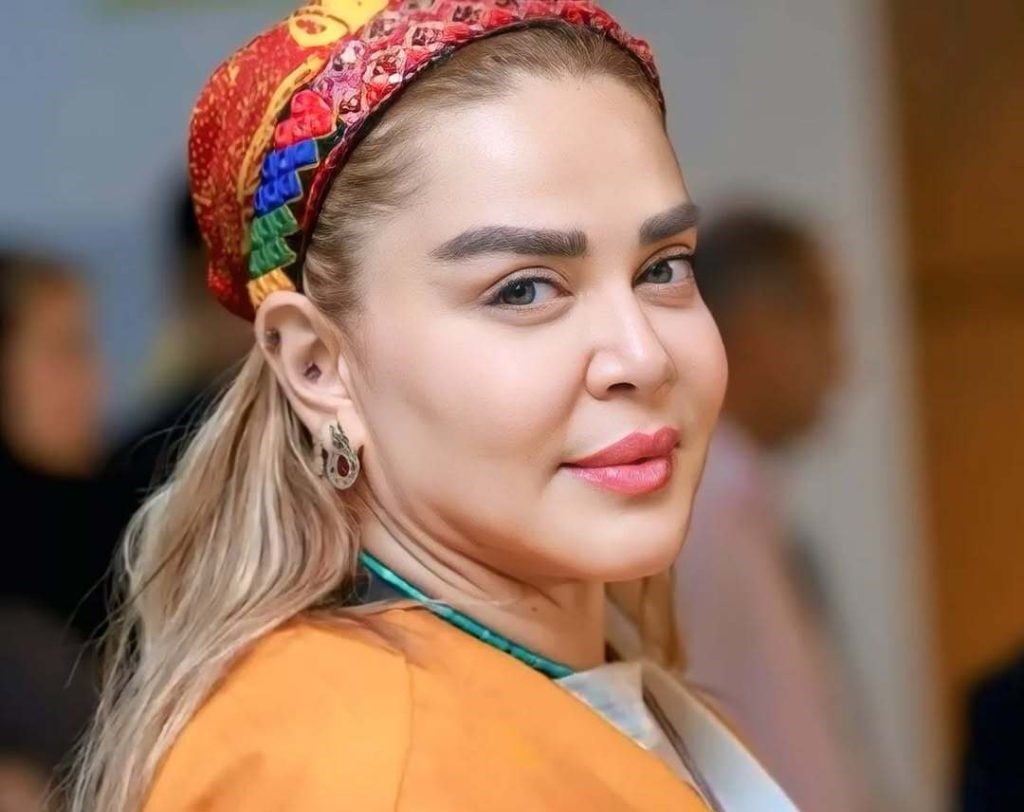 Portrait of a Smiling Woman with Blonde Hair Wearing a Colorful Patterned Headscarf and Orange Top Facing the Camera with Earrings Visible