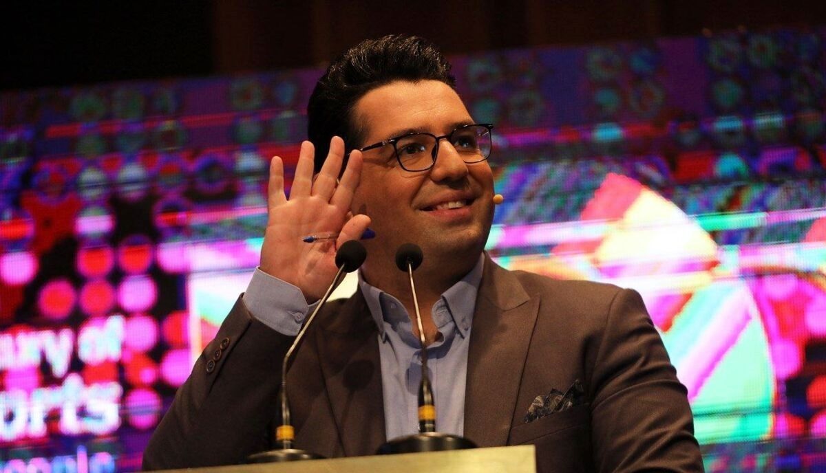 Speaker at a Podium Gesturing with His Left Hand While Addressing an Audience Two Microphones in Front of Him Colorful Stage Backdrop Behind Him