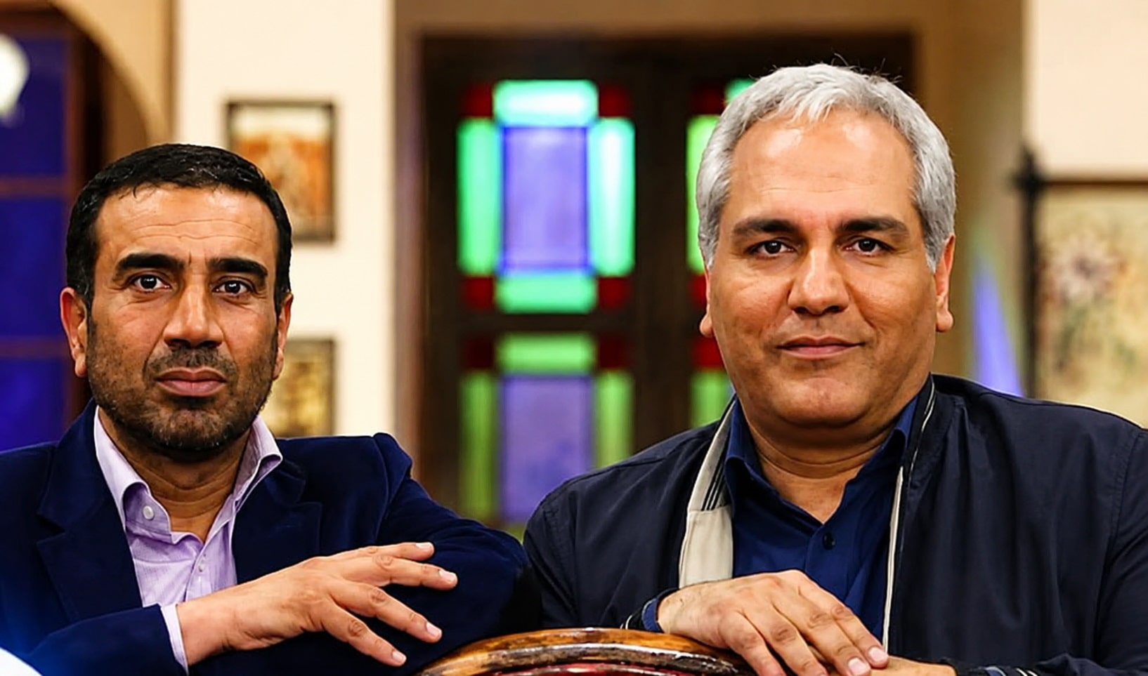 Two Men Pose for a Photo Indoors Leaning on a Wooden Table with a Colorful Stained glass Window Behind Them