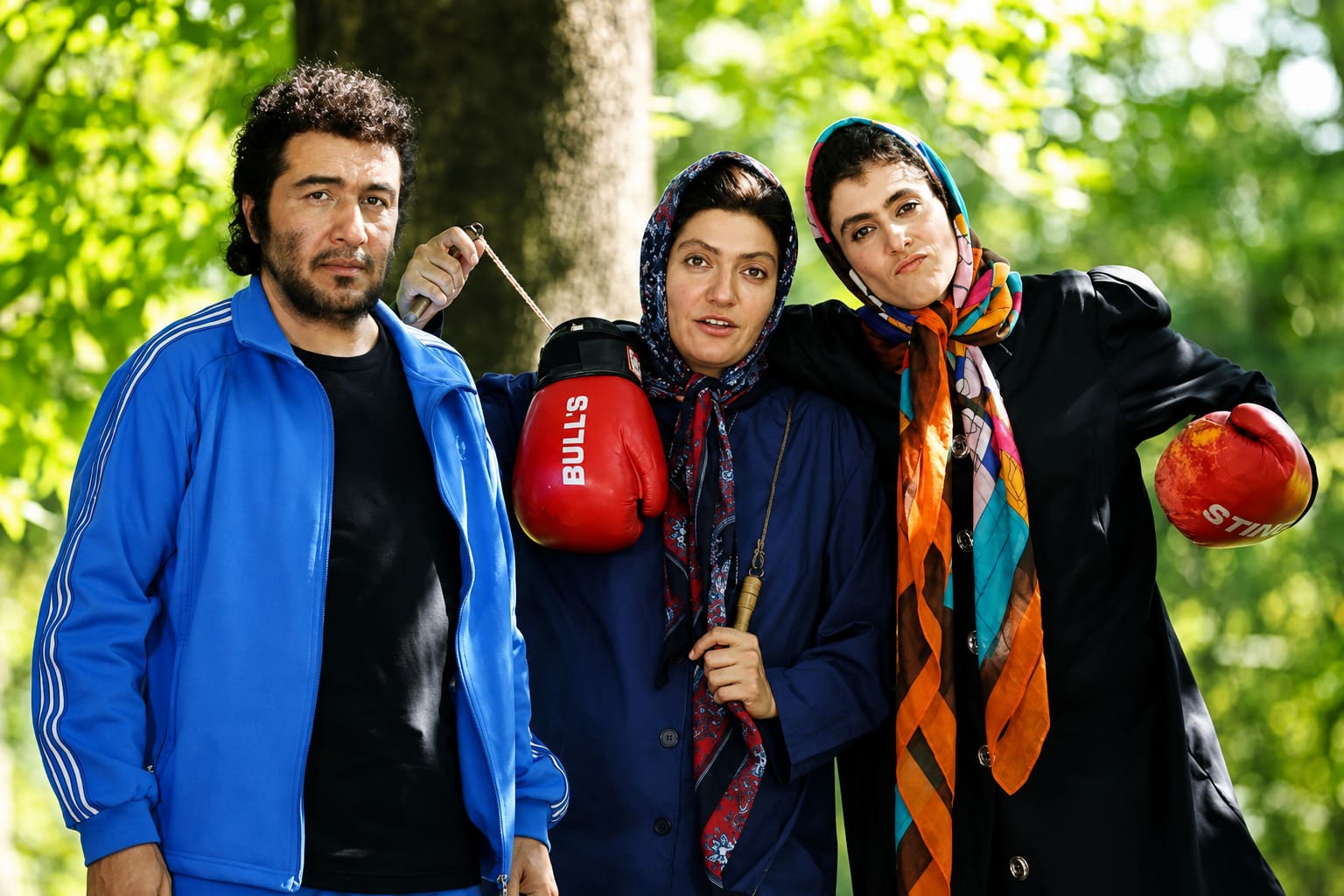 Three Friends Posing in a Sunny Park Two Women Wearing Headscarves Hold Red Boxing Gloves While a Man in a Blue Jacket Stands Beside Them
