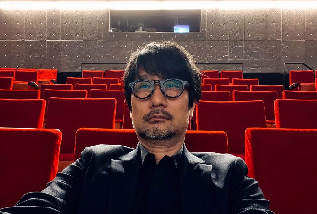 Self portrait of a Man Wearing Round Glasses and a Dark Suit Seated in an Empty Red Theater Auditorium