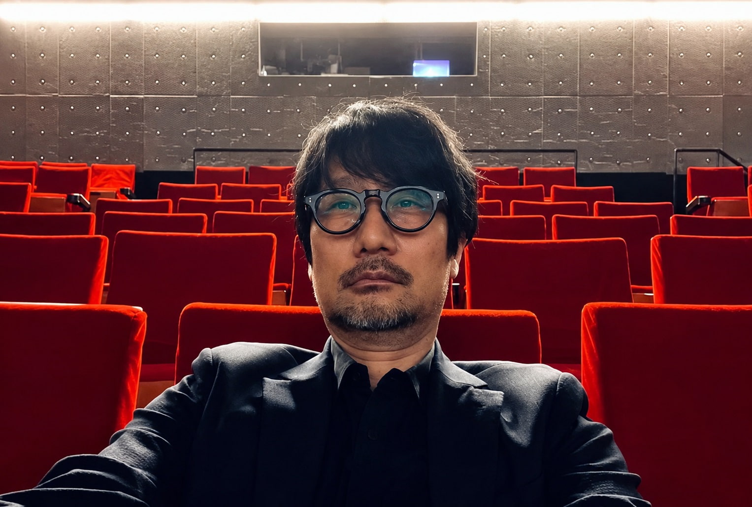 Self portrait of a Man Wearing Round Glasses and a Dark Suit Seated in an Empty Red Theater Auditorium