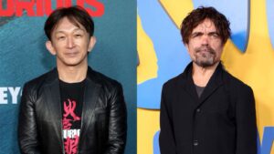 Two Men Posing at a Red carpet Event Left Man Wears a Black Leather Jacket and Graphic Tee Right Man Wears a Dark Blazer Before a Blueyellow Backdrop