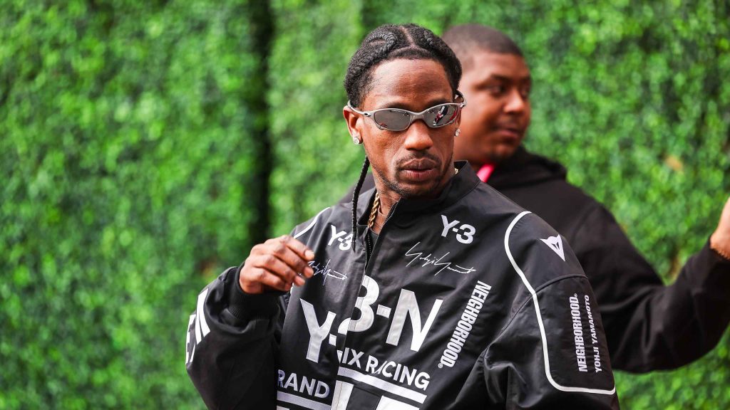 Man Wearing a Black Racing Jacket with White Lettering and Mirrored Sunglasses Outdoors in Front of a Green Hedge