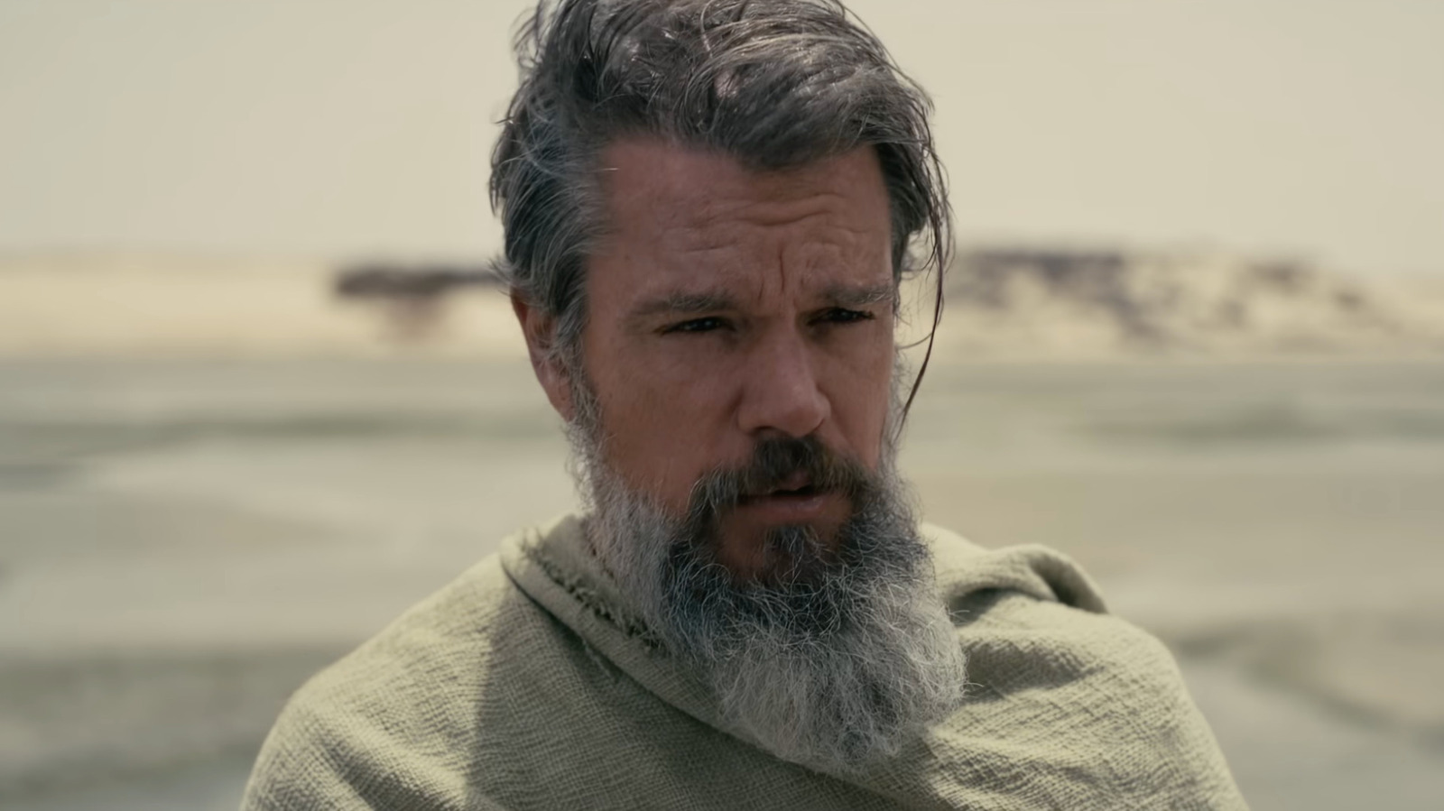 Man with Gray Beard Wrapped in a Rough Beige Cloak Stands on a Windswept Beach Gazing off to the Side with a Serious Expression